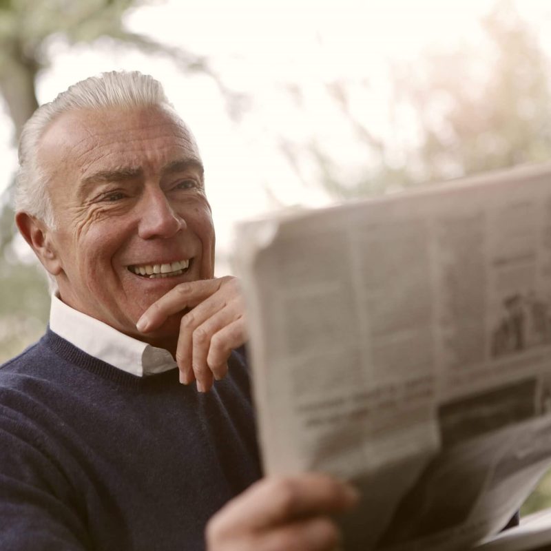 Man smiling while reading a newspaper outdoors.